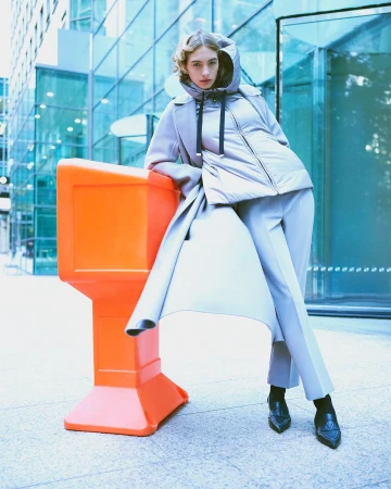 Fashionable woman in winter outerwear poses outdoors in city against a modern building, leaning against an orange traffic barrier.