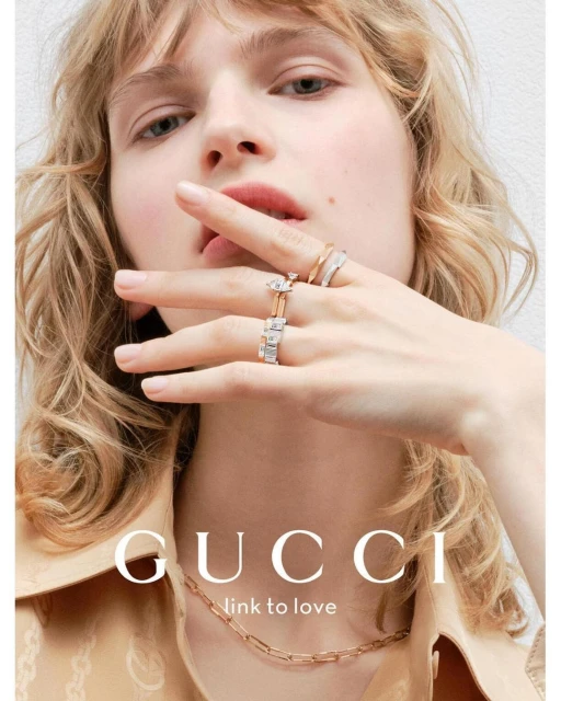 Close-up of a blonde female model showcasing Gucci jewelry, including rings and a necklace, in a studio with soft lighting.