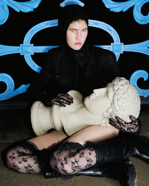Fashion shot of a young woman with pale skin, wearing dark clothing and sitting on a pedestal, hugging a bust outdoors.