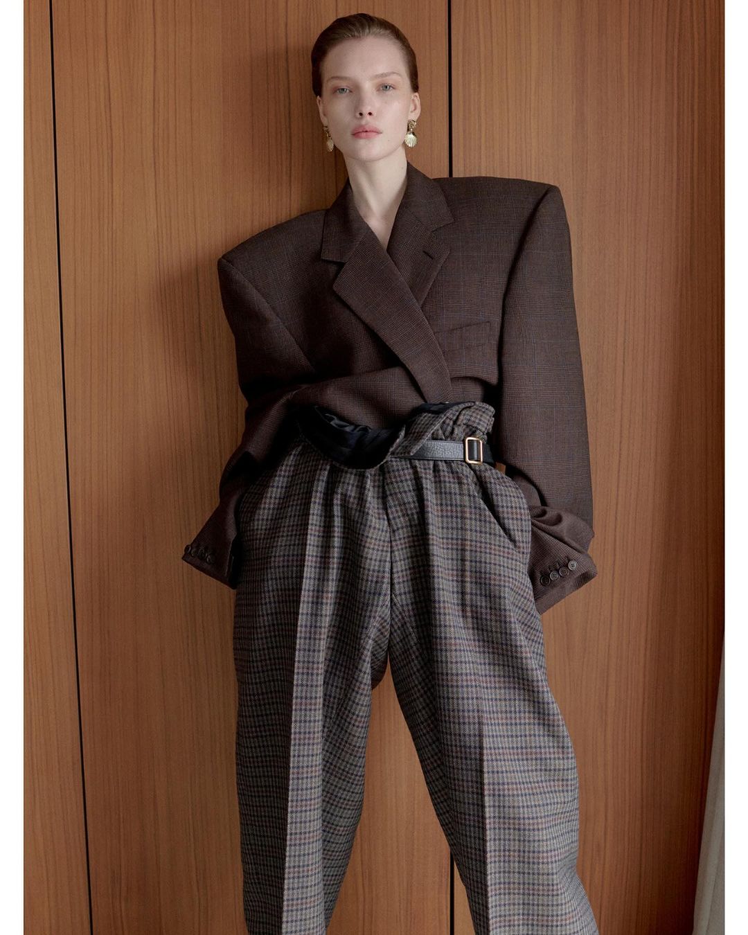 Woman in oversized brown blazer and plaid trousers posing against wood paneling in a studio. Fashion editorial portrait.