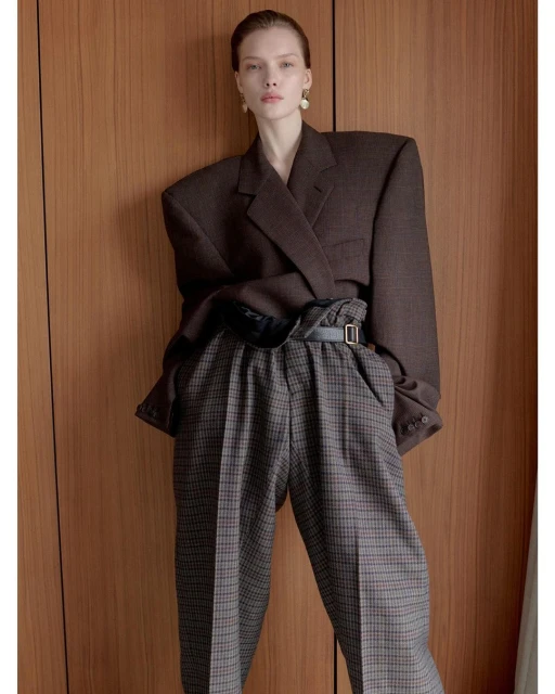 Woman in oversized brown blazer and plaid trousers posing against wood paneling in a studio. Fashion editorial portrait. 