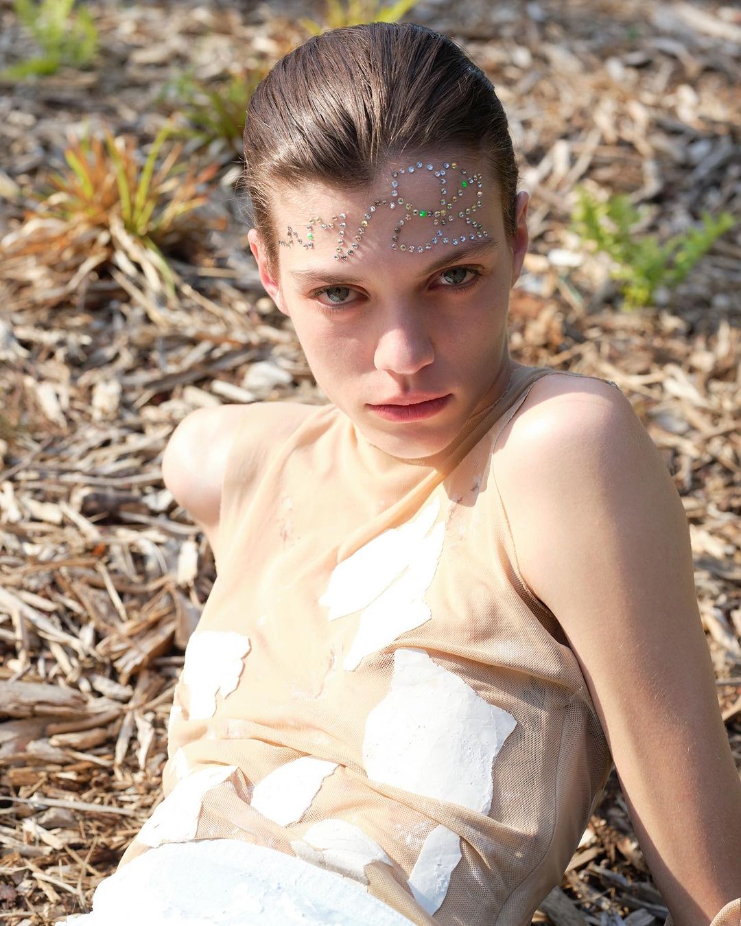 A young woman with fair skin, wearing a light top with embellishments, poses outdoors in natural light on a wood-chip surface.