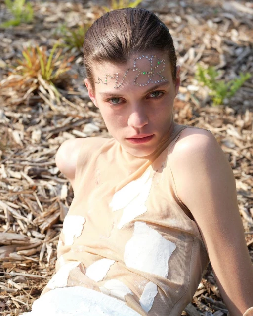 A young woman with fair skin, wearing a light top with embellishments, poses outdoors in natural light on a wood-chip surface.