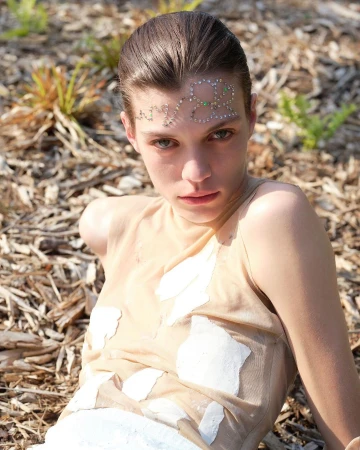 A young woman with fair skin, wearing a light top with embellishments, poses outdoors in natural light on a wood-chip surface.