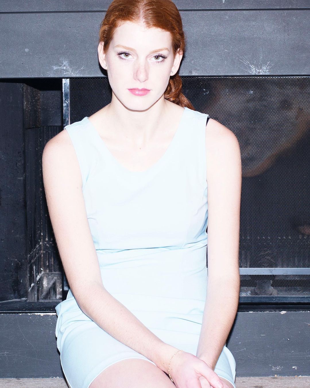 Red-haired woman wearing a pale blue sleeveless dress seated indoors in front of a dark fireplace.