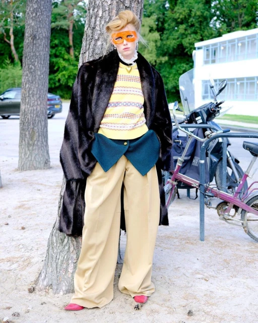 Fashionable woman in wide-leg pants and fur coat poses outdoors near trees and a bicycle, wearing bright orange glasses.