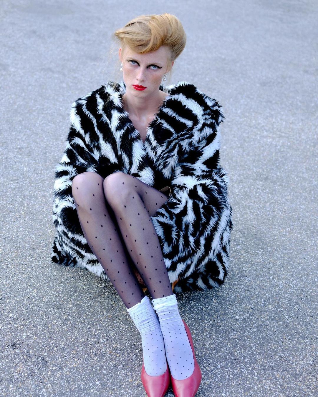 Stylish adult female model with blond updo, in spotted tights, pink shoes, and a zebra-print fur coat, sitting outdoors on concrete.