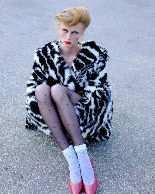 Stylish adult female model with blond updo, in spotted tights, pink shoes, and a zebra-print fur coat, sitting outdoors on concrete.