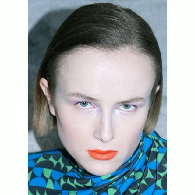 Close-up portrait of a model with bold orange lips and lilac eyeshadow wearing a patterned blue, green, and black top against a grey backdrop.