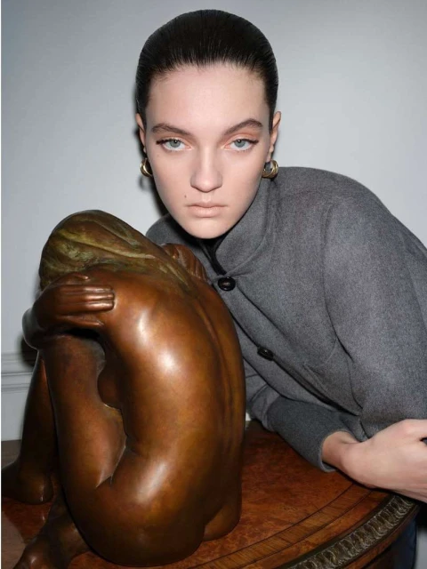 Female model poses in gray coat next to bronze sculpture of a crouching nude woman in a studio with soft, diffused lighting.