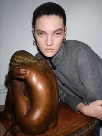 Female model poses in gray coat next to bronze sculpture of a crouching nude woman in a studio with soft, diffused lighting.