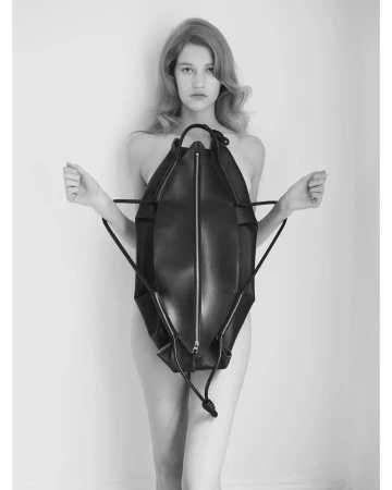 Black and white studio shot of a woman with light hair holding a large, dark backpack against a light wall backdrop, fashion editorial.