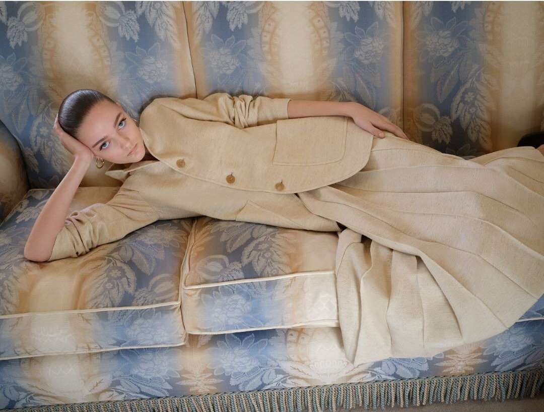 Woman in a beige jacket and pleated skirt lying on a floral patterned couch with a fringe, indoor setting, fashion photography.