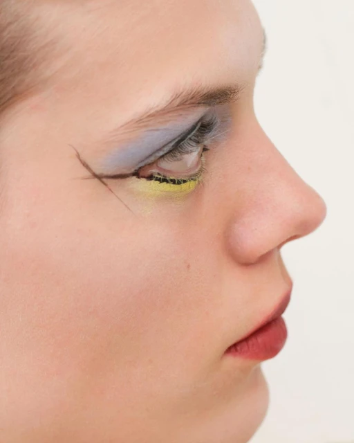 Close-up of a young woman's face in profile, featuring dramatic eye makeup with gray eyeshadow and yellow eyeliner, set against a white background.