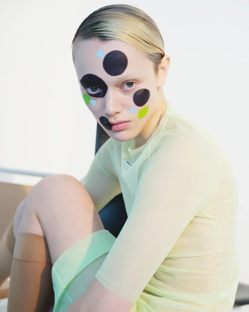 Portrait of a young woman with dots painted on her face, wearing a sheer yellow shirt and seated indoors against a neutral background.