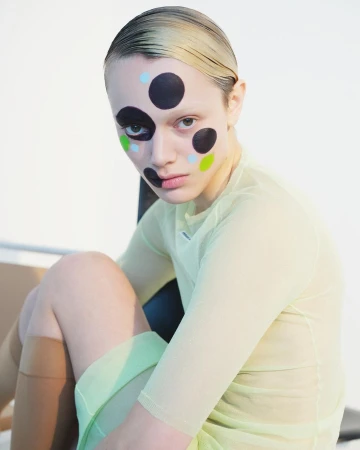 Portrait of a young woman with dots painted on her face, wearing a sheer yellow shirt and seated indoors against a neutral background.