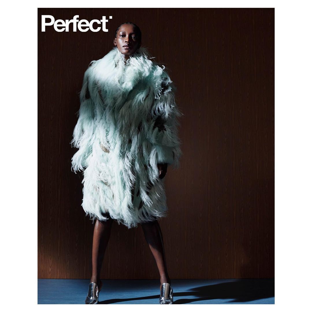 Fashion editorial shot of a Black woman in a textured teal coat and silver heels, posing against a brown backdrop with magazine text.