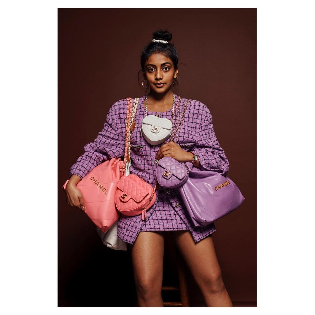 Woman in tweed skirt suit poses indoors with a collection of Chanel handbags in pastel colors against a brown backdrop. Fashion shoot.