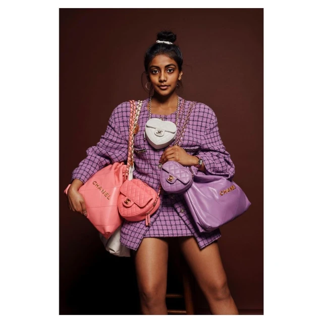 Woman in tweed skirt suit poses indoors with a collection of Chanel handbags in pastel colors against a brown backdrop. Fashion shoot.