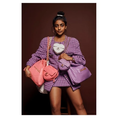 Woman in tweed skirt suit poses indoors with a collection of Chanel handbags in pastel colors against a brown backdrop. Fashion shoot.