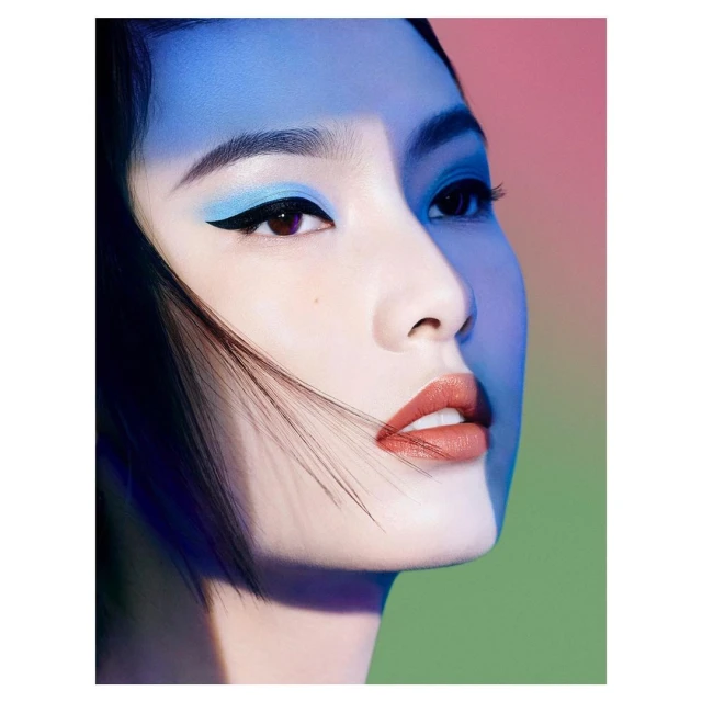 Close-up beauty shot of a woman with bold makeup including blue eyeshadow, black eyeliner, and coral lipstick against a colorful backdrop.