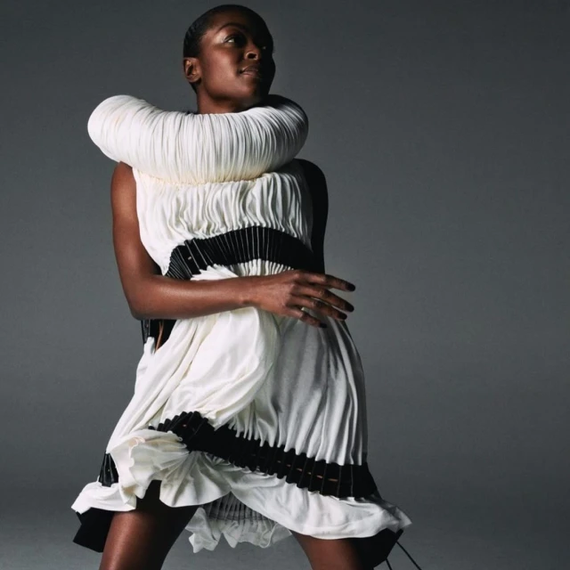 Woman poses in a unique white sculptural dress with black accents against a grey studio backdrop, highlighting fashion and portrait photography.