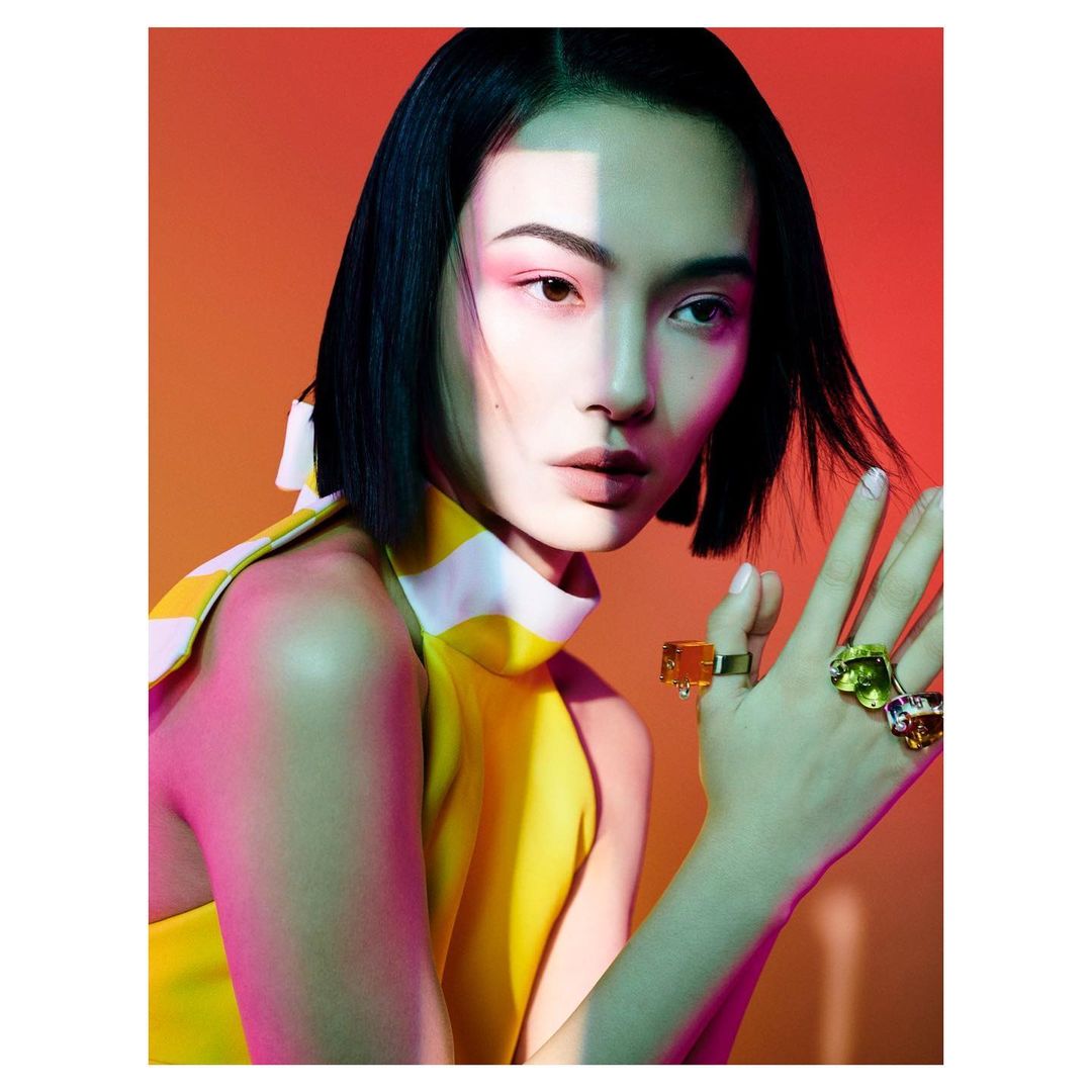 Glamour shot of Asian woman with bob haircut wearing yellow halter top with large gemstone rings, posing in front of orange background.