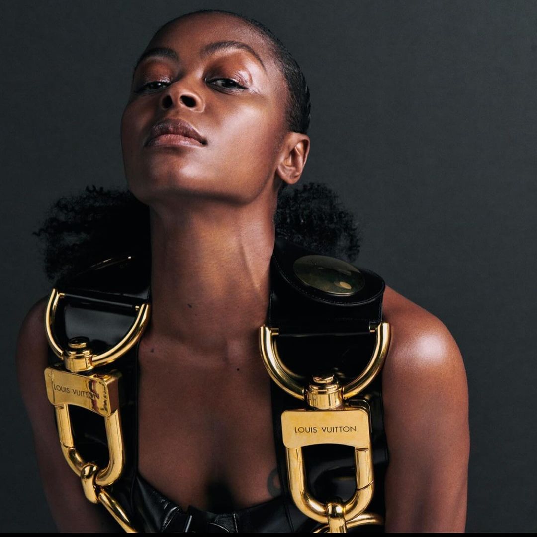 Adult woman with dark skin wearing a black top and gold-buckled harness in a studio shot against a dark background.