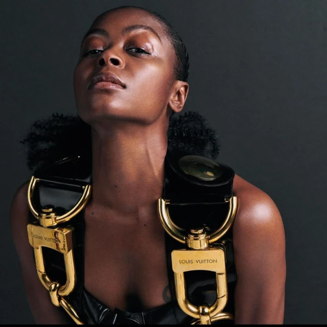 Adult woman with dark skin wearing a black top and gold-buckled harness in a studio shot against a dark background.