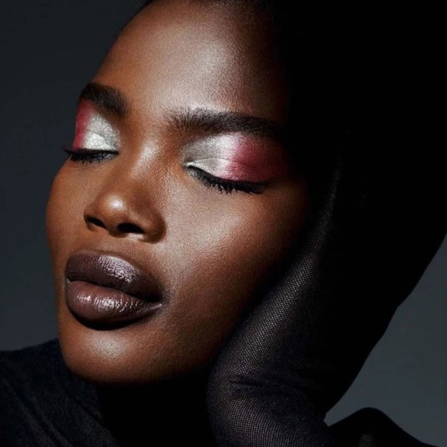 Close-up portrait of a dark-skinned woman with closed eyes, wearing dramatic silver and red eyeshadow, and featuring glossy dark lips.