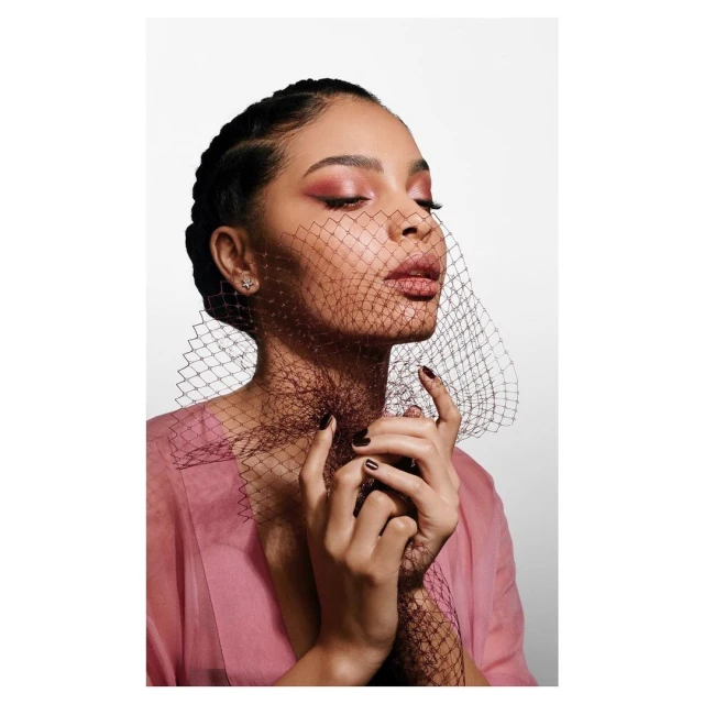 Close-up portrait of a woman with her eyes closed, wearing a pink top and black mesh veil in front of a solid white background.