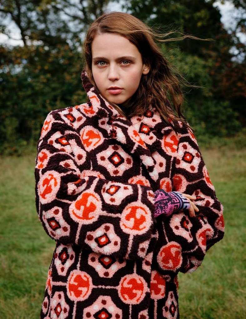 A young, fair-skinned woman with auburn hair poses outdoors in a patterned coat, looking directly at the camera with a neutral expression.