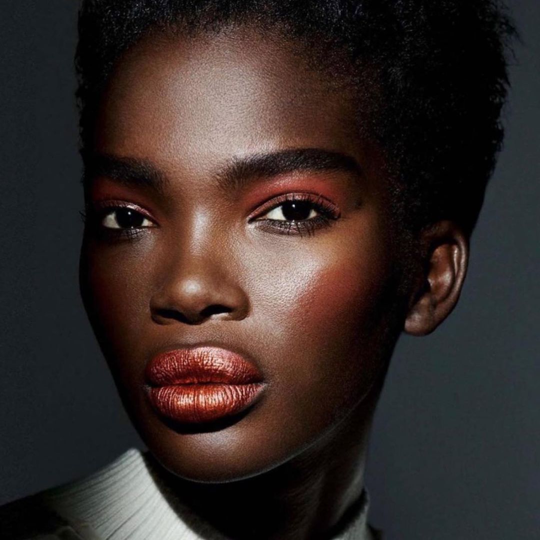 Close-up studio portrait of a dark-skinned African woman, with short hair and copper makeup on the eyelids and lips, against a dark gray background.