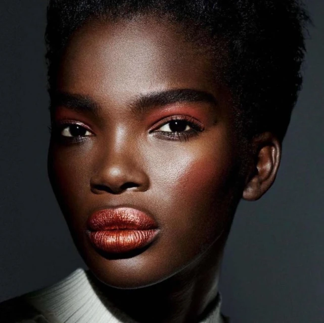 Close-up studio portrait of a dark-skinned African woman, with short hair and copper makeup on the eyelids and lips, against a dark gray background.