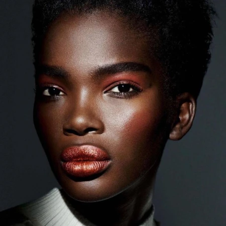 Close-up studio portrait of a dark-skinned African woman, with short hair and copper makeup on the eyelids and lips, against a dark gray background.