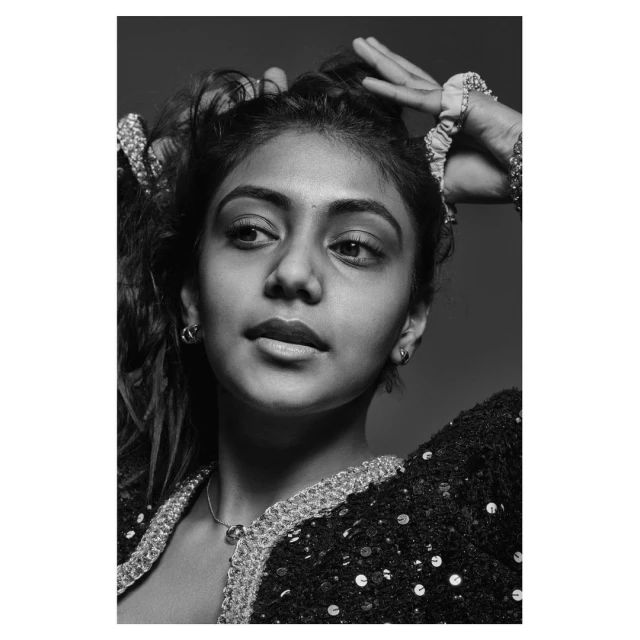 Monochrome portrait of a woman with dark hair, fair skin, hands in hair, wearing a dark sequined jacket and simple jewelry, studio lighting.
