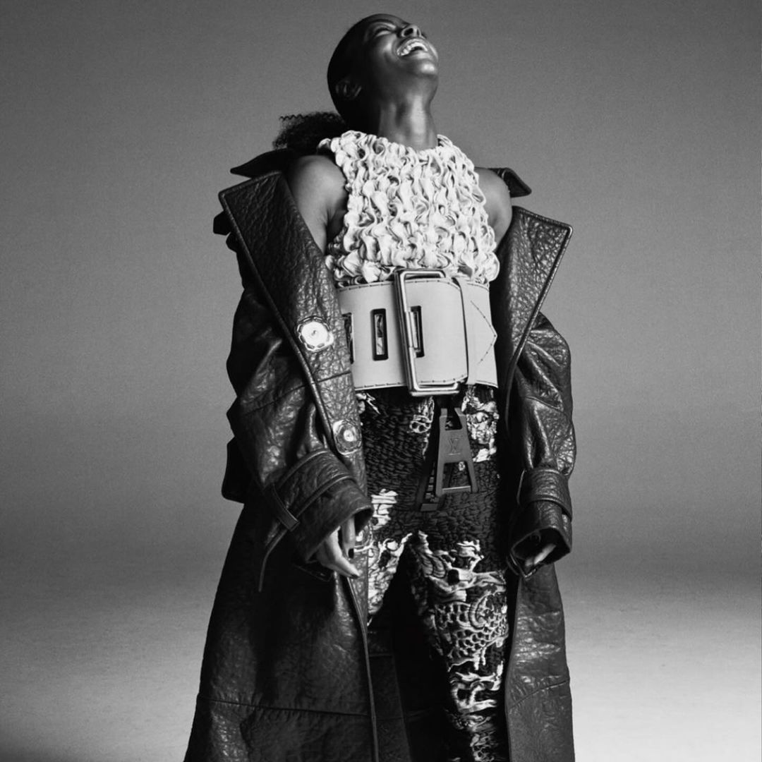 A black and white fashion portrait of a woman with short hair wearing a textured top, wide belt, patterned pants, and a leather jacket.