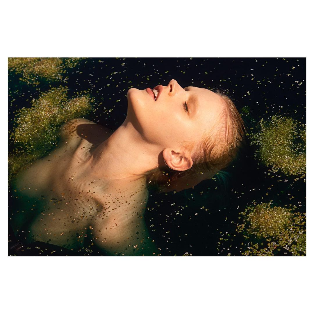 Woman in a pond with water plants. Her head is tilted back, and her eyes are closed. Water droplets are visible on her skin.