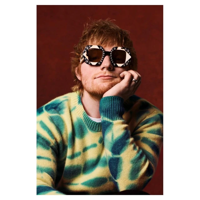 Portrait of singer Ed Sheeran with a beard, wearing patterned glasses and a tie-dye sweater, against a red background in a studio.