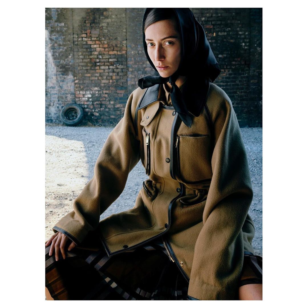 Woman in a tan coat and black headscarf poses against a brick wall. Neutral colors and natural light create an editorial fashion image.
