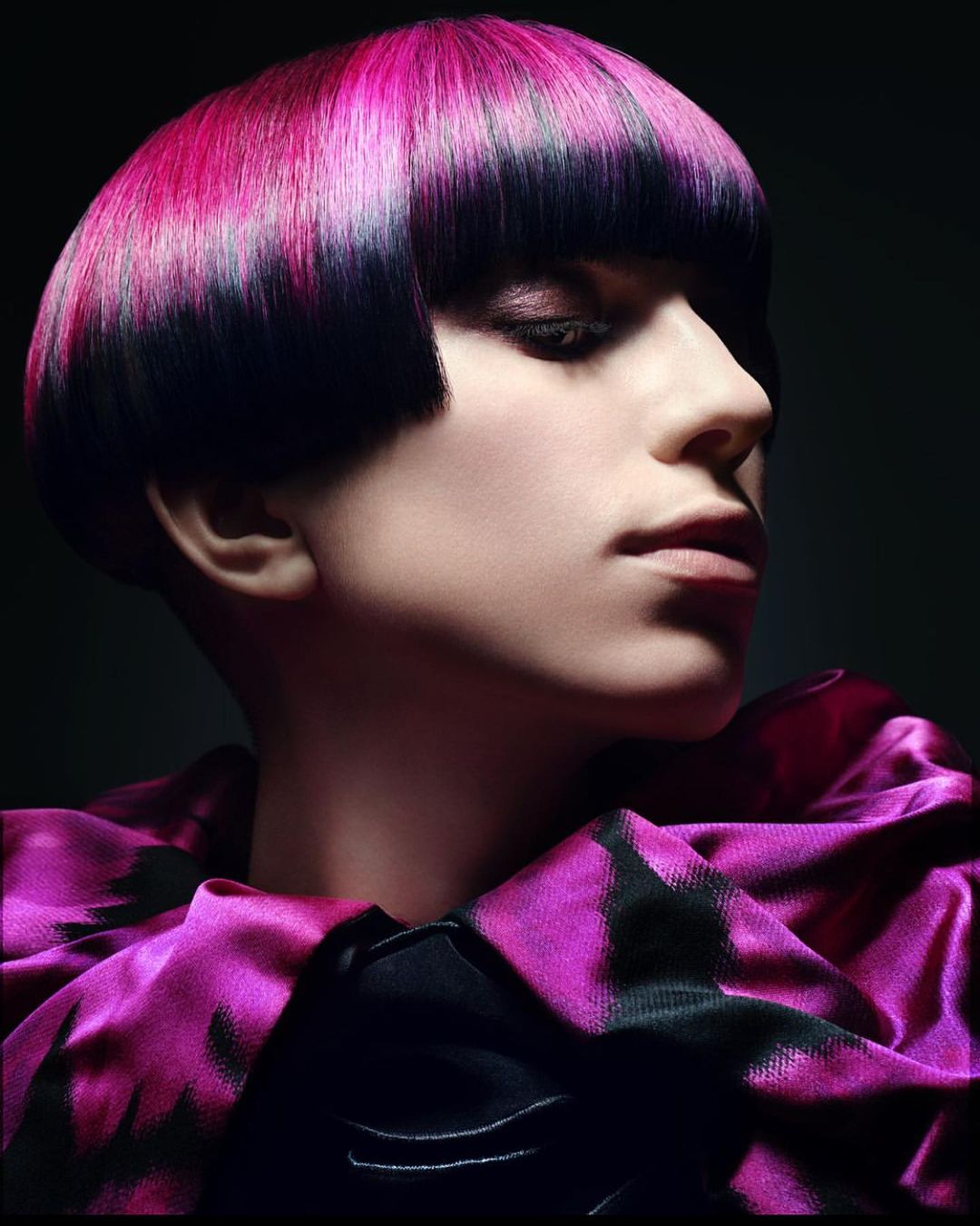 Close-up of a woman with a stylish bowl haircut dyed black and pink, wearing a dark pink patterned top against a dark background.