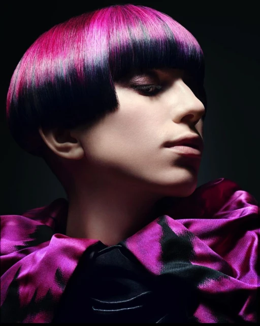 Close-up of a woman with a stylish bowl haircut dyed black and pink, wearing a dark pink patterned top against a dark background.