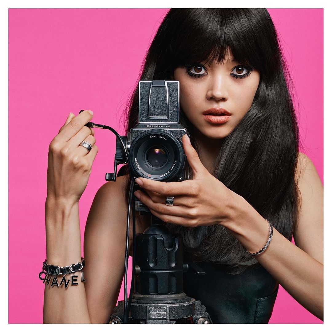 Portrait of a woman holding a vintage camera on a tripod against a bright pink backdrop, showcasing fashion bracelets and rings.