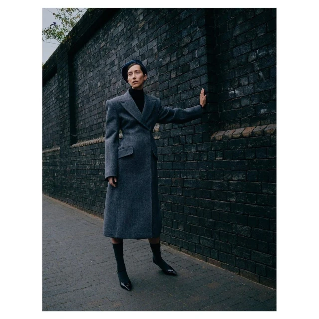 A stylish adult female stands in a long grey wool coat and black sock boots against a dark brick wall in an urban setting.