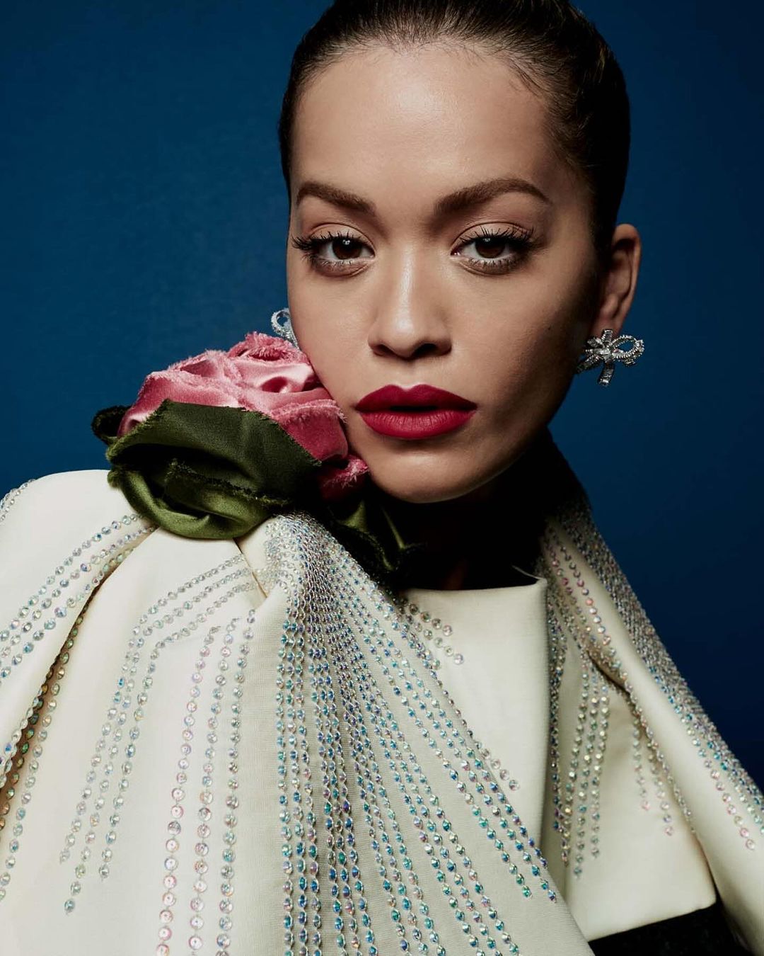 Close-up portrait of a woman with red lipstick wearing a rhinestone-studded cape with a rose on her shoulder against a blue backdrop.