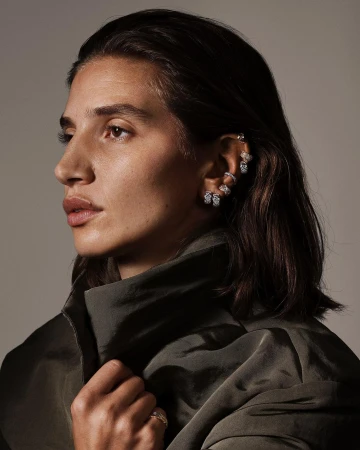 Profile portrait of a woman wearing diamond ear cuffs and a dark olive high-collar jacket against a grey background.
