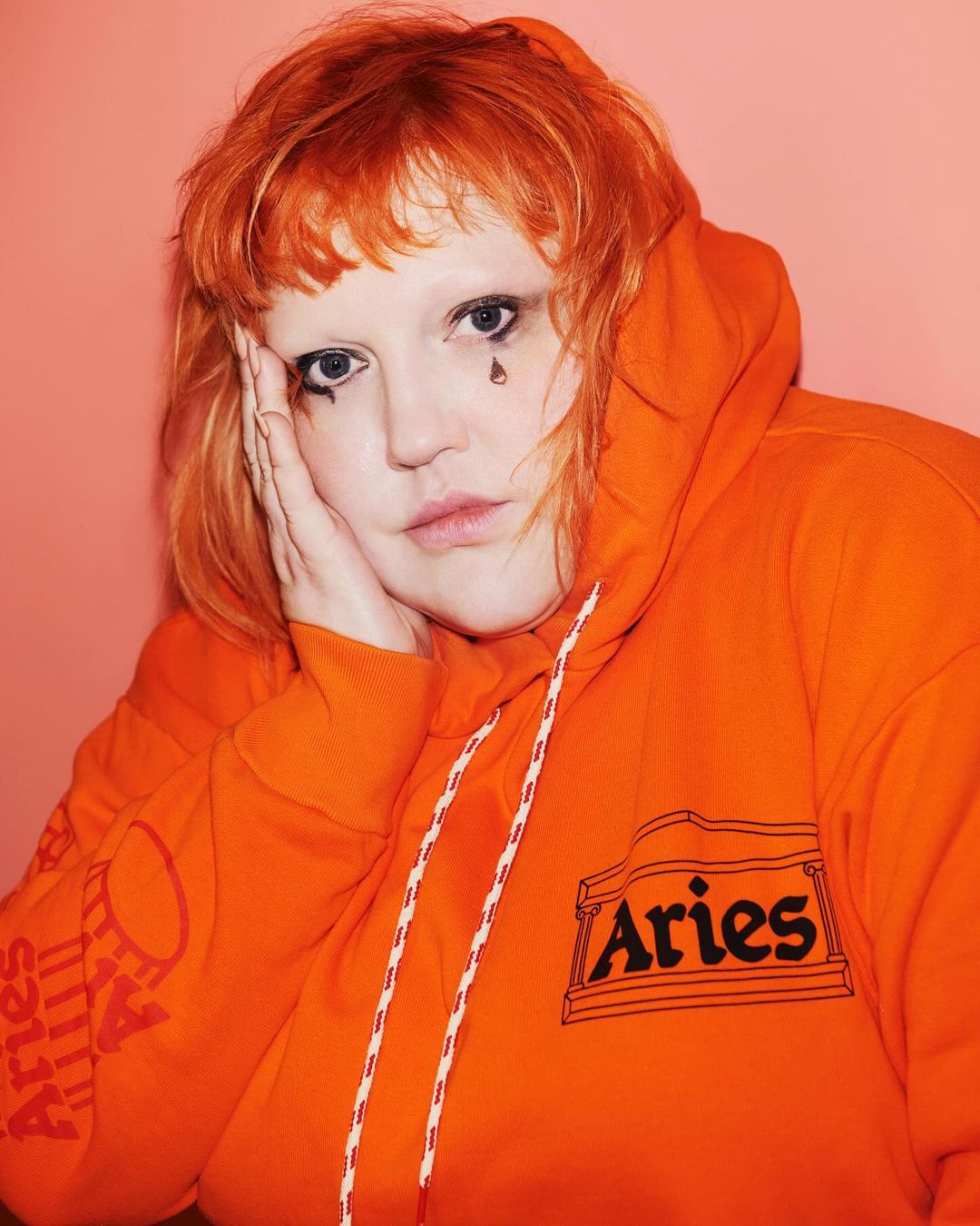 Close-up portrait of a red-haired adult female wearing an orange hoodie and tear-shaped makeup, set against a pink background.