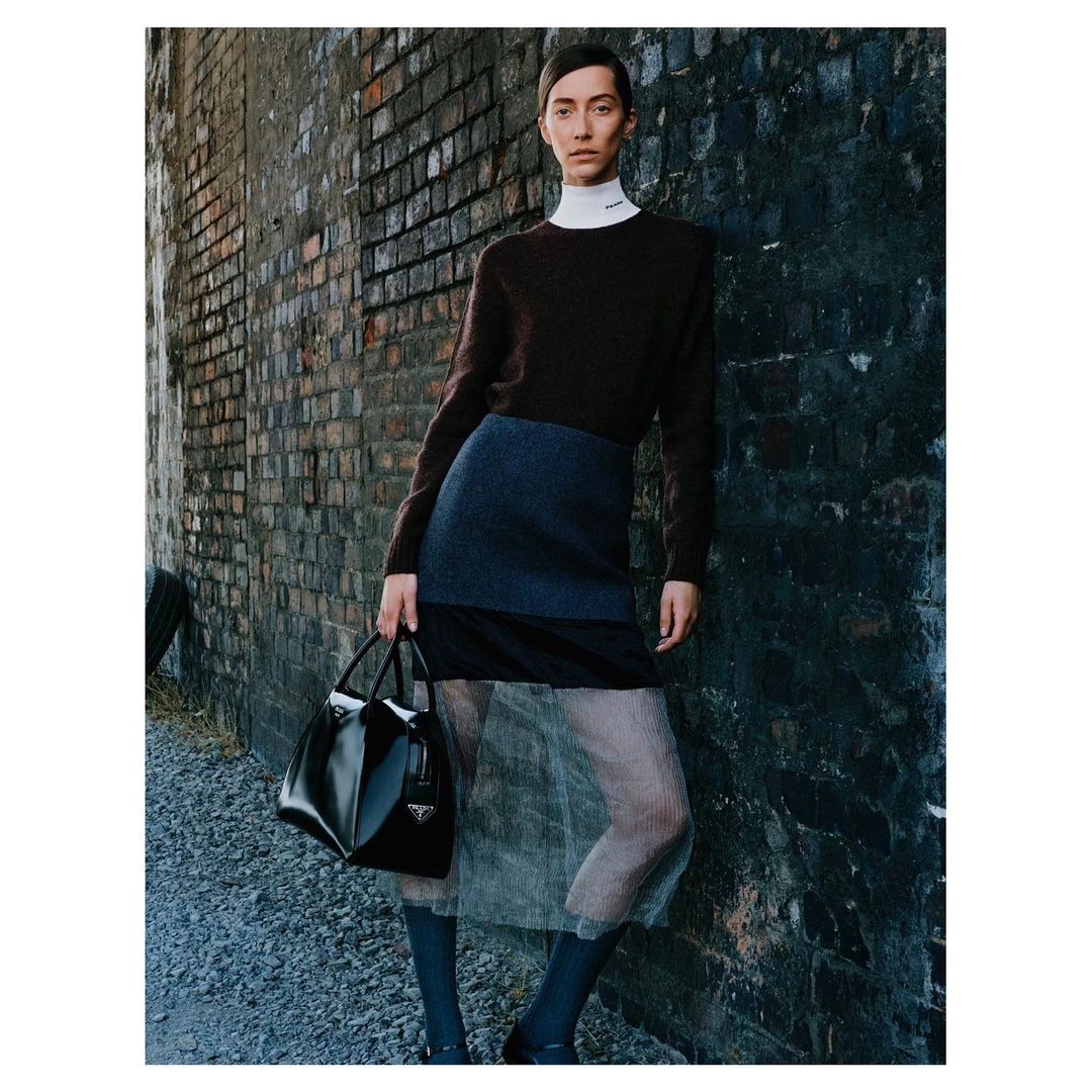 A slender fashion model leans against a brick wall, holding a black Prada purse, wearing a sweater, skirt, sheer tights, and dark socks.