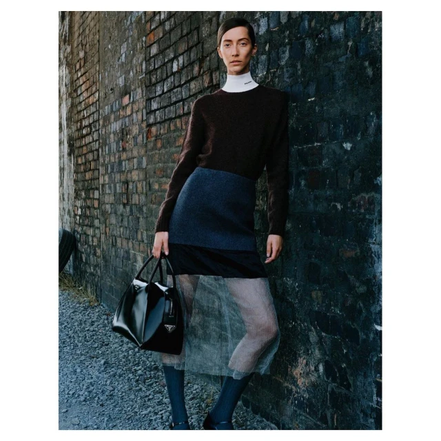 A slender fashion model leans against a brick wall, holding a black Prada purse, wearing a sweater, skirt, sheer tights, and dark socks.