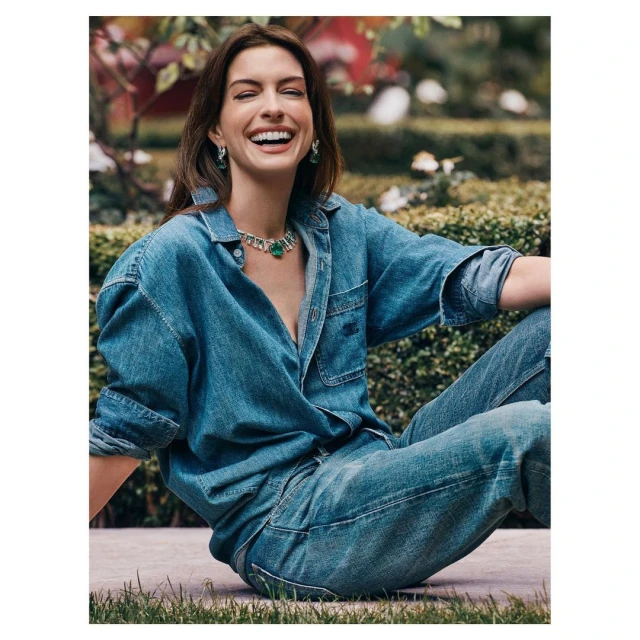 Anne Hathaway sits outside in a denim shirt and jeans, laughing and wearing jewelry, with a natural background of greenery.
